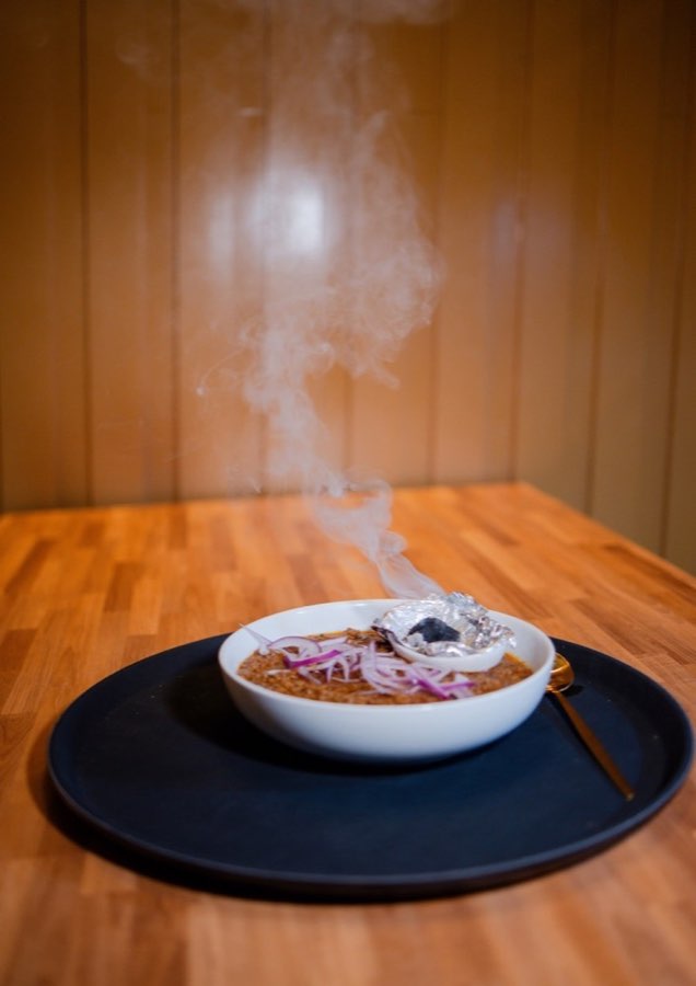 Alvi's smoked haleem topped with sliced red onion in a white bowl, with smoke rising from a small charcoal cup.