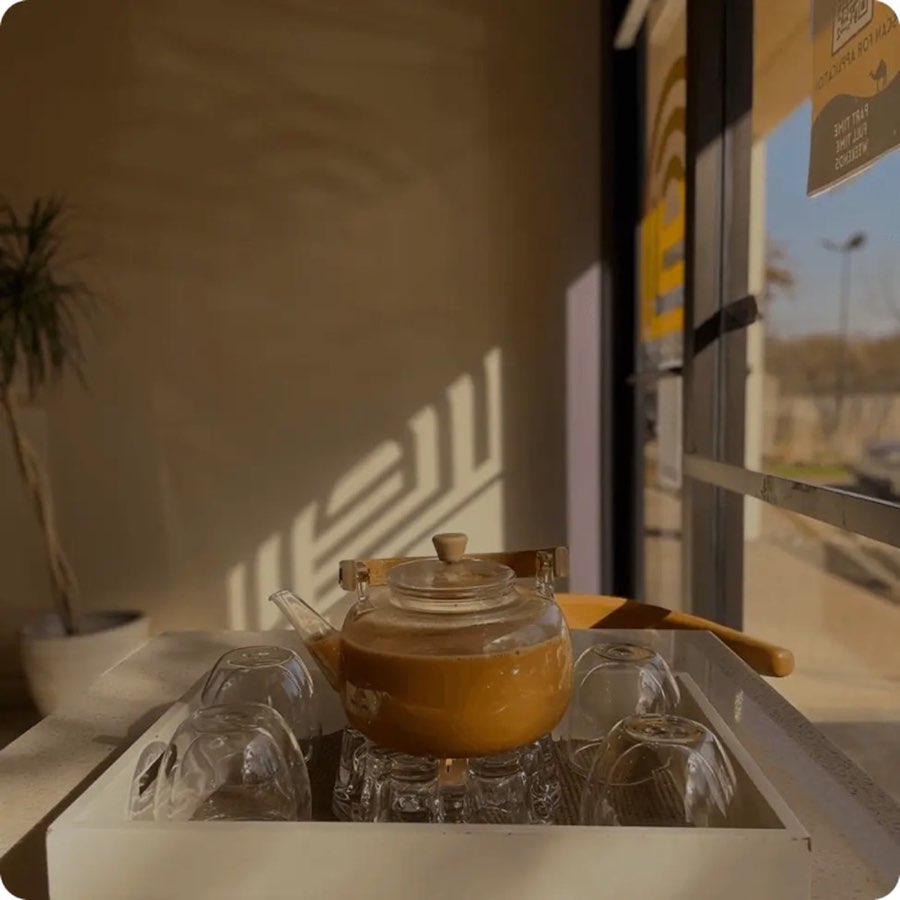 Glass pot of a milky hot drink with small serving glasses on a tray by an Arwa café window