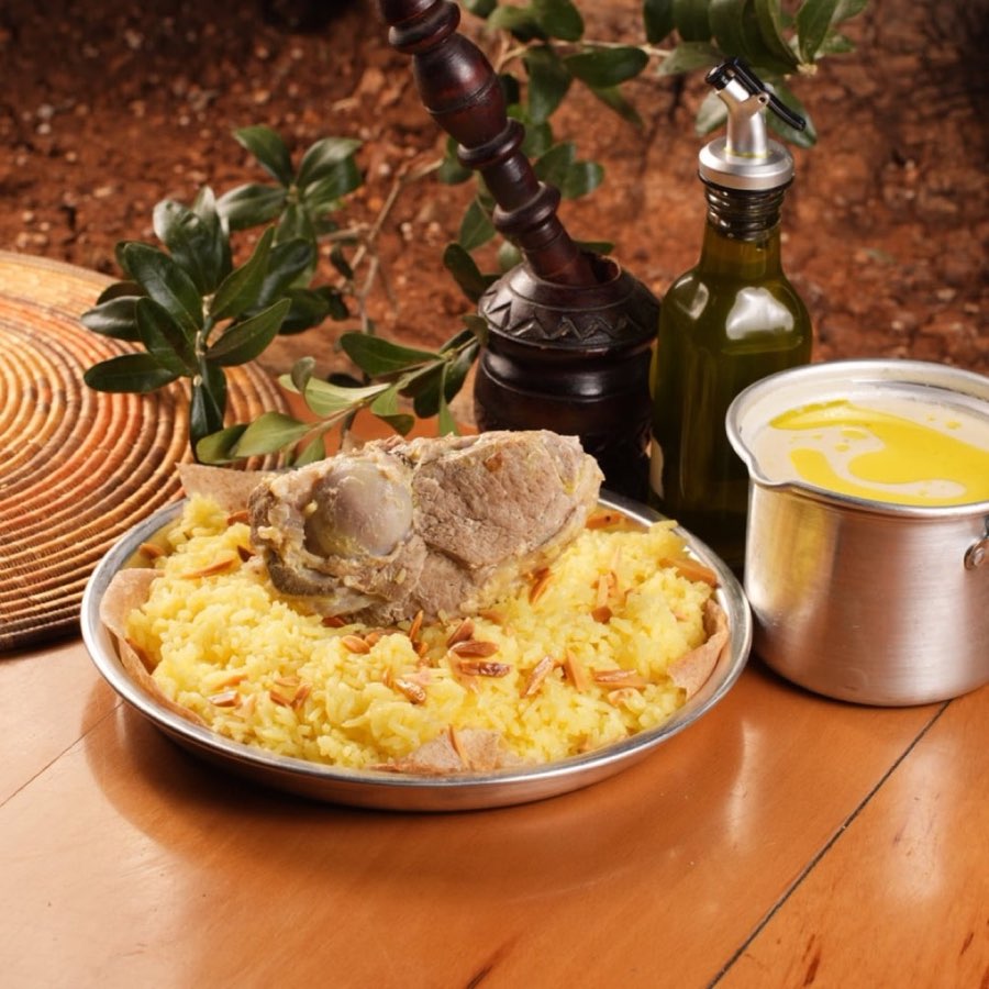Khashoka Jordanian mansaf: a lamb portion over yellow rice and nuts, with a pot of pale jameed sauce on a wooden table.